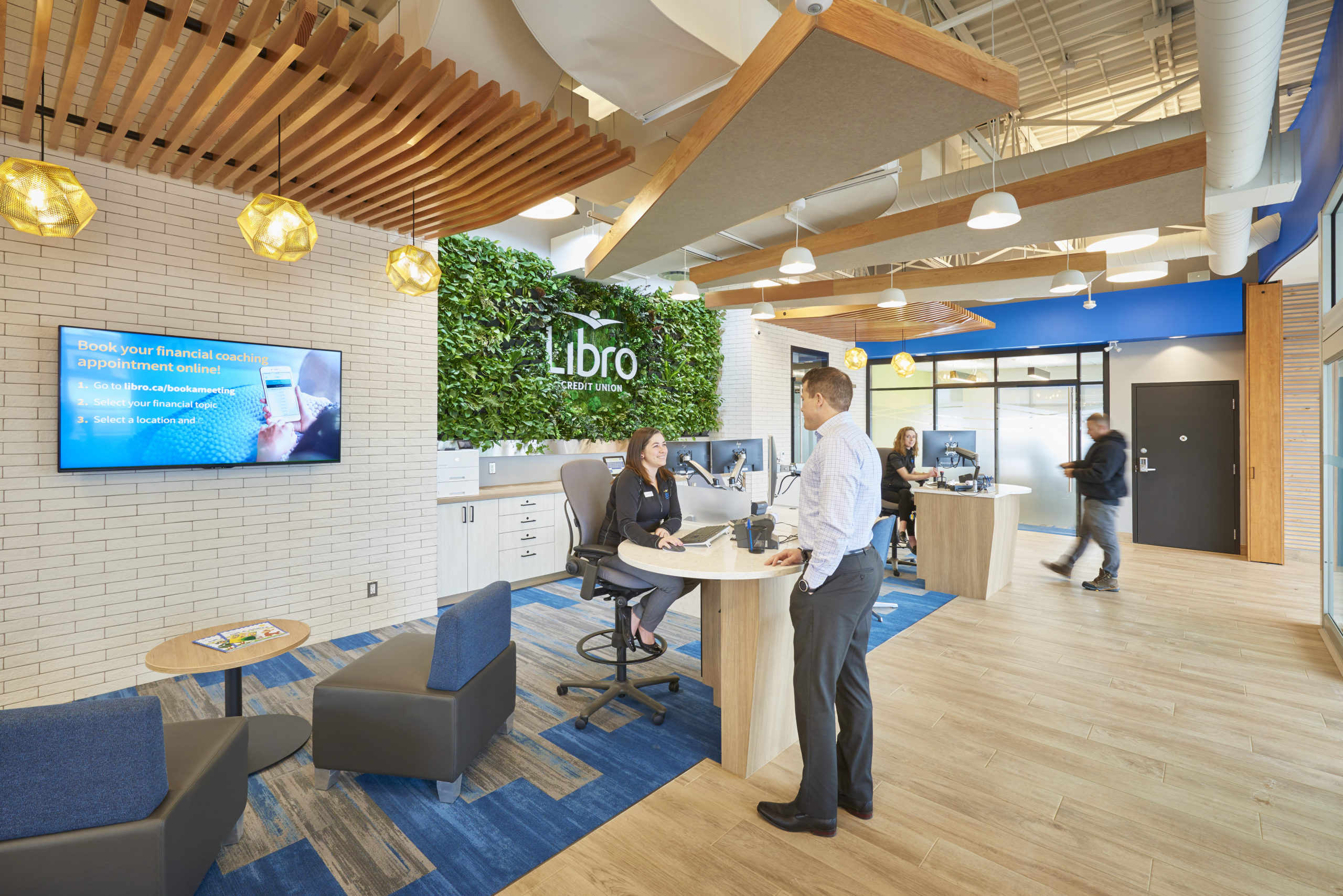 interior of Libro Credit Union lobby technology screen on the wall over a seating area, man standing at a counter speaking to a woman