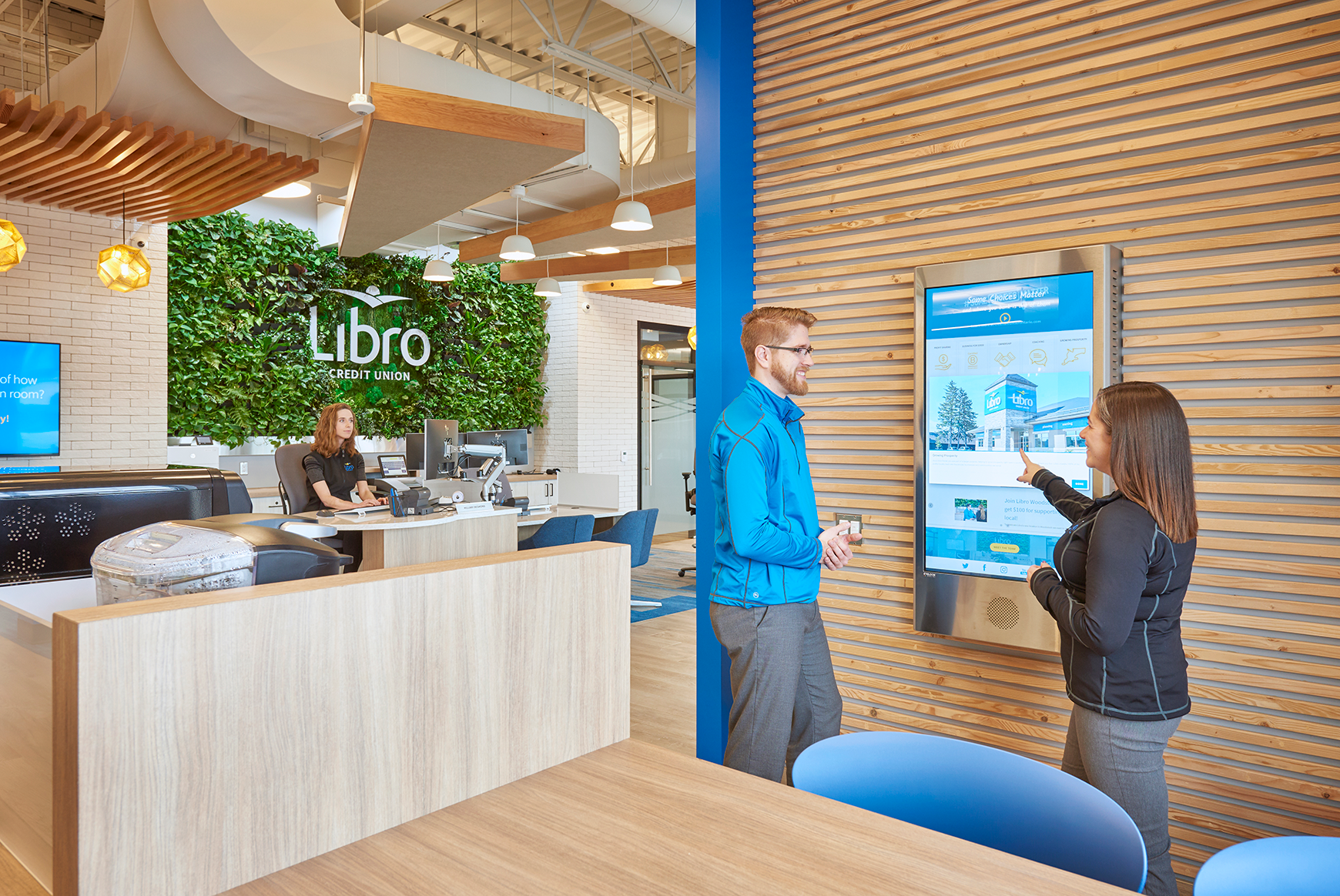 a woman sitting at a desk working and two people having a discussion around a screen in the Libro Credit Union lobby