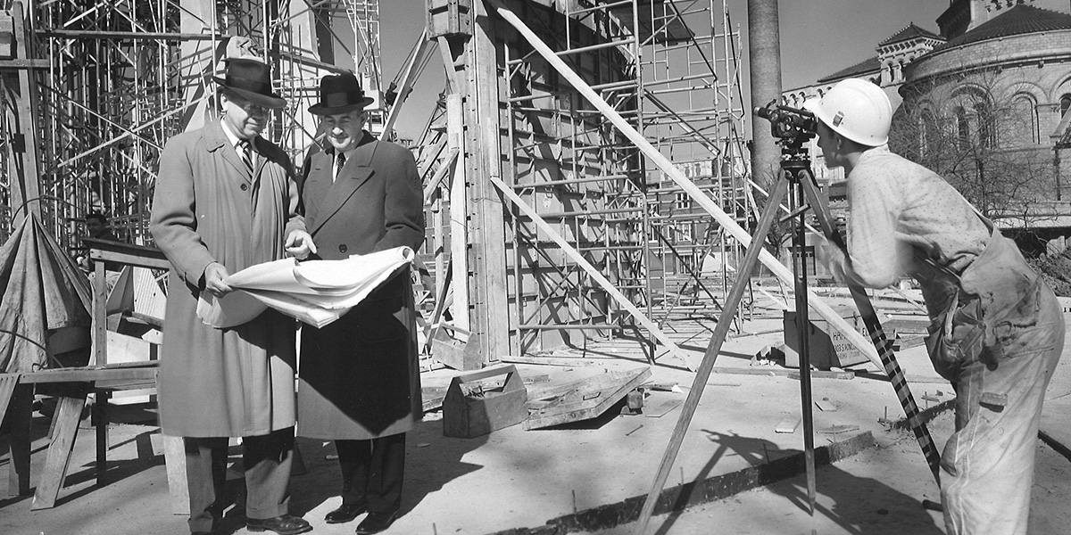 black and white photo from the history of Newground two men in long coats and hats looking at blueprints